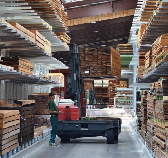 Operator verplaatst hout dat hoog is opgeslagen op draagarmstellingen met een elektrische pallettruck
		                    